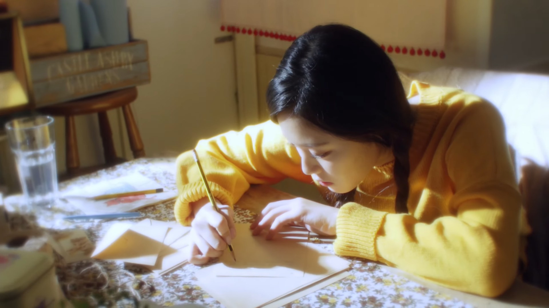 Hyunjin sitting at a table and writing on a piece of paper. There are other pieces of paper strewn across the table.